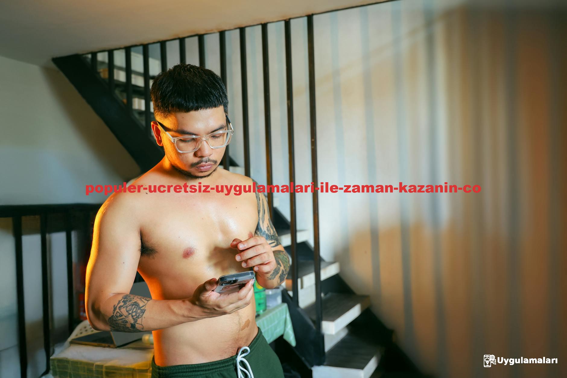 Shirtless young man with tattoos using smartphone by indoor staircase, showcasing casual lifestyle.